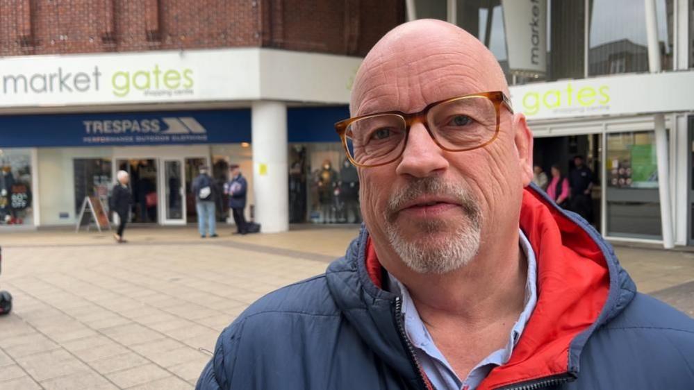 Jonathan Newman, wearing a blue shirt and jacket with red hood lining. He is bald and has a greying goatee beard and is wearing glasses. He is standing near the Great Yarmouth Market Gates Shopping Centre.