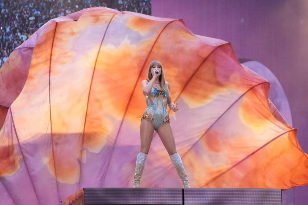 Taylor Swift standing on stage singing, holding a microphone up to her face. She is wearing a sparkly blue and gold bodysuit and sparkly silver boots. She is stood against a background of pink, purple and orange material, with some of the crowd visible behind.