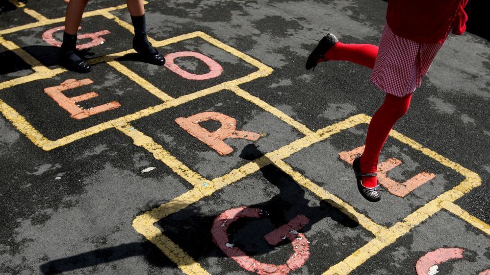 A stock photo shows two children playing hopscotch in a school playground.