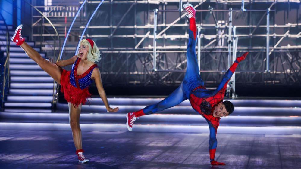 Woman with blonde hair wearing a blue top and red skirt with one leg up in the air. Beside her is a man with short dark hair in a blue and red Spiderman outfit using one hand to hold himself up with his legs in the air. They are both on a dancefloor with scaffolding and a set of stair in the background. 