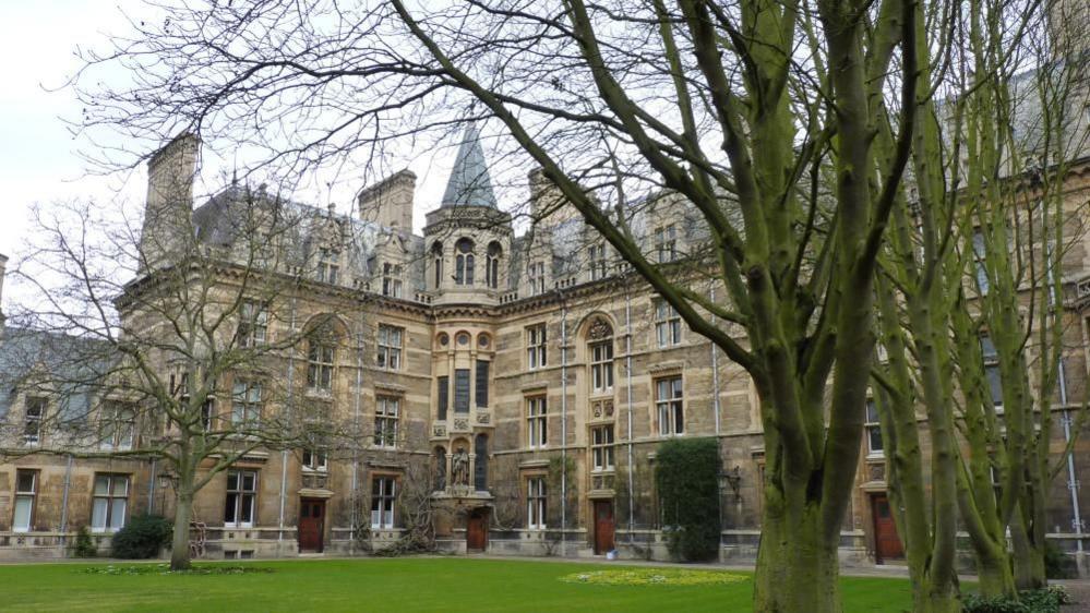 An ornate Cambridge college building. It is made of sand coloured bricks and sits behind a lawn.