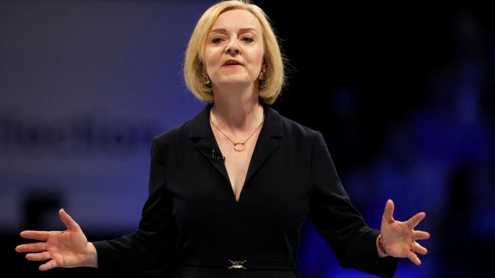 Liz Truss in black jacket holding arms out