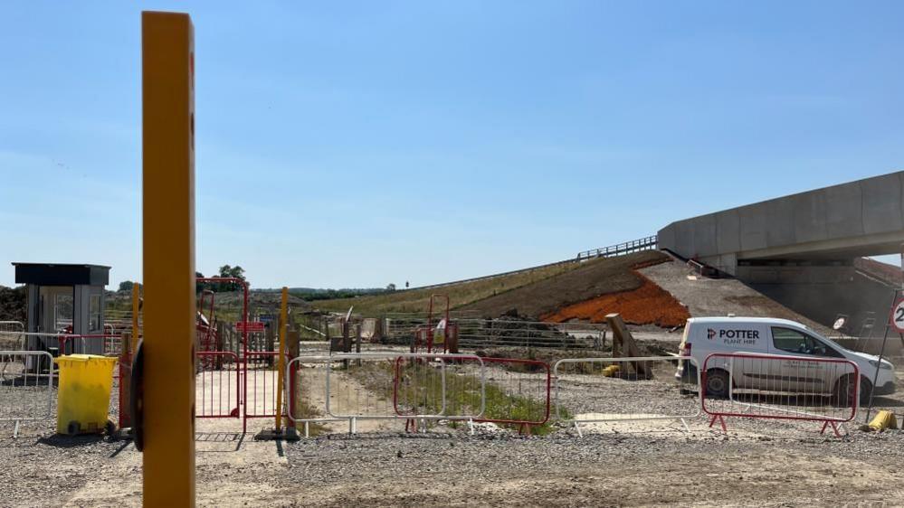 A white van parked in a temporary car park surfaced with stones. There is a red railing with a gate in the foreground. Behind the car park is a newly-hewn hill leading to a stone overbridge.