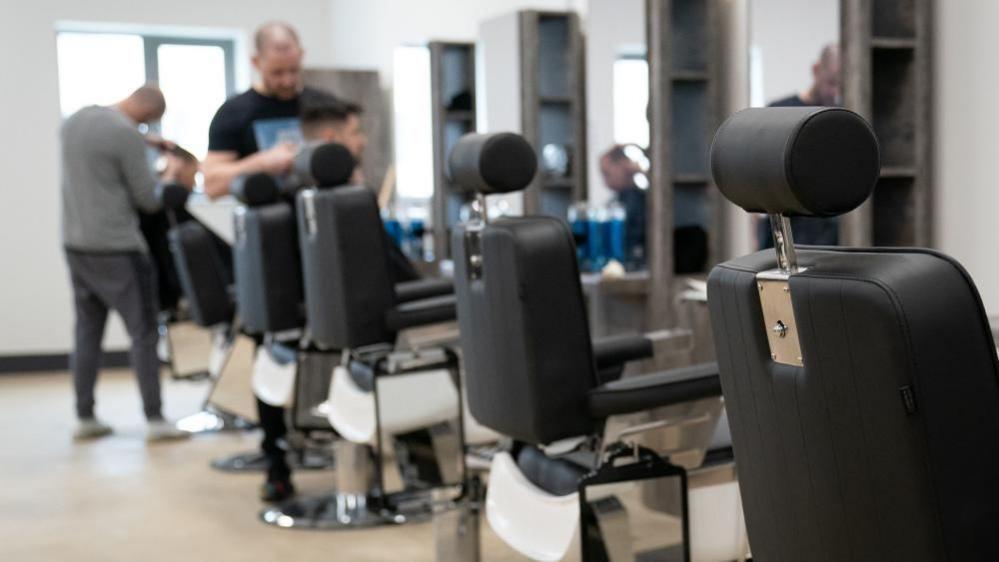 Row of clack barber's chairs with headrests and mirrors opposite. Two people are standing up cutting hair of "customers" whose faces are blurred.