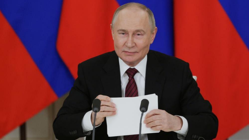 Vladimir Putin. The Russian president is seated in front of a Russian flag and behind two microphones and is holding a piece of paper as he makes an address. He is wearing a dark suit, white shirt and red tie