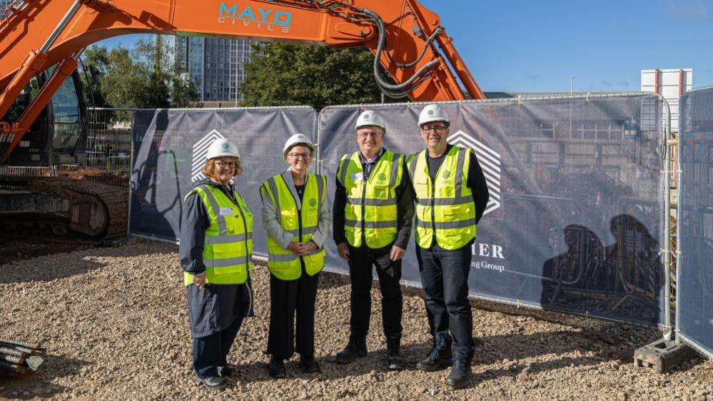 Two women and two men, one of whom is Andy Burnham, stand wearing high-vis vests and white hard hats on a building site. An excavator is parked behind them.