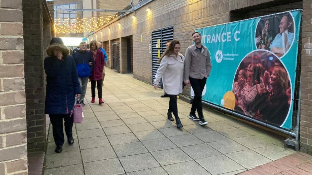 Large Entrance C poster on the wall as people walk by on grey paving stones.