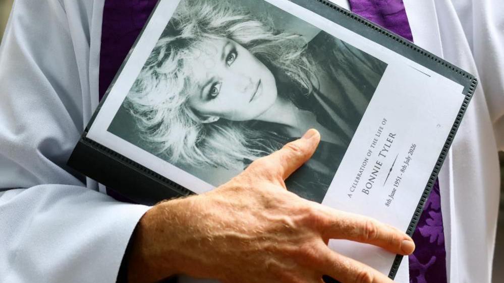 A priest's hand and part of their white robe with a purple patterned stole can be seen holding an order of service with a black and white photo of Bonnie Tyler and 'A celebration of the life of Bonnie Tyler' and '8th June 1951- 8th July 2026' written on the front.