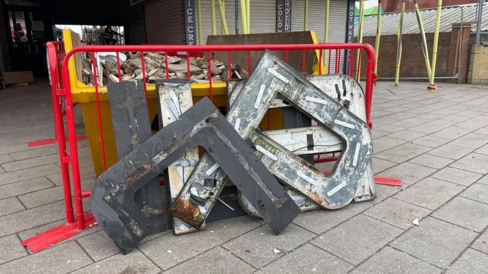 Metal letter backing plates, next to a red barrier and a skip outside the Britannia Pier.