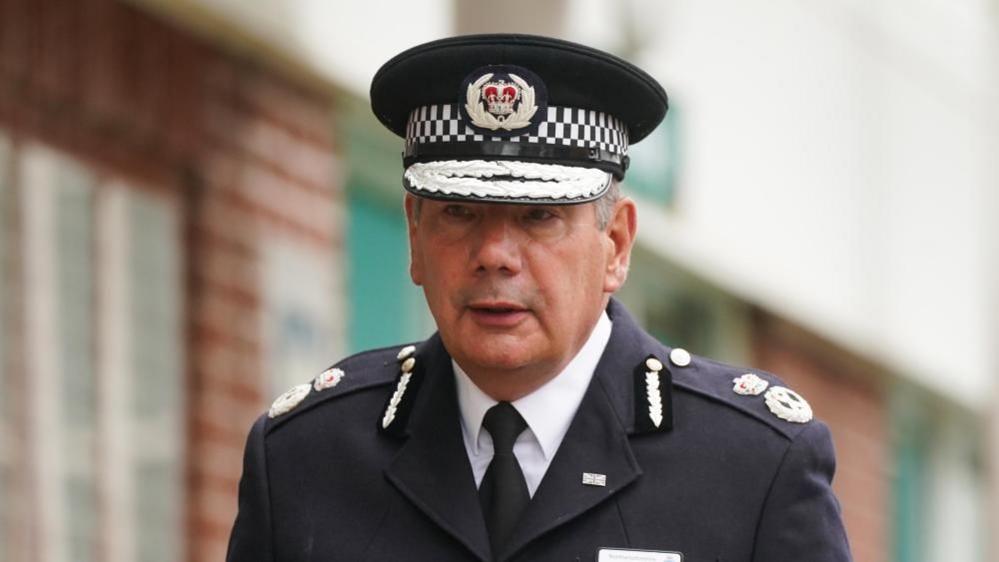 Nick Adderley with short, dark hair wearing a police cap, black jacket and black tie. He is outside a brick-built building.