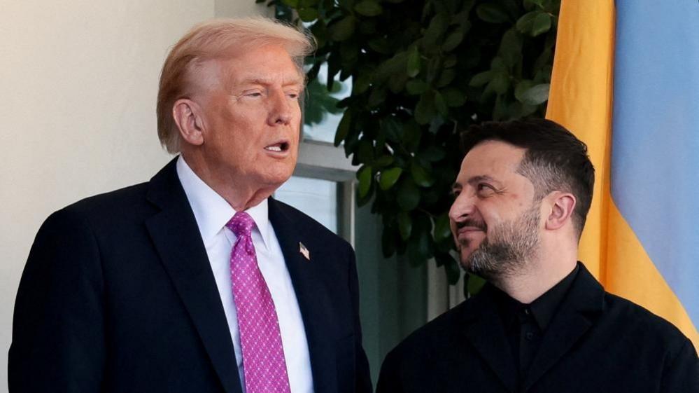 Donald Trump, wearing a white short and chequered violet and white tie speaks as Volodymyr Zelensky all in black looks up to him smiling