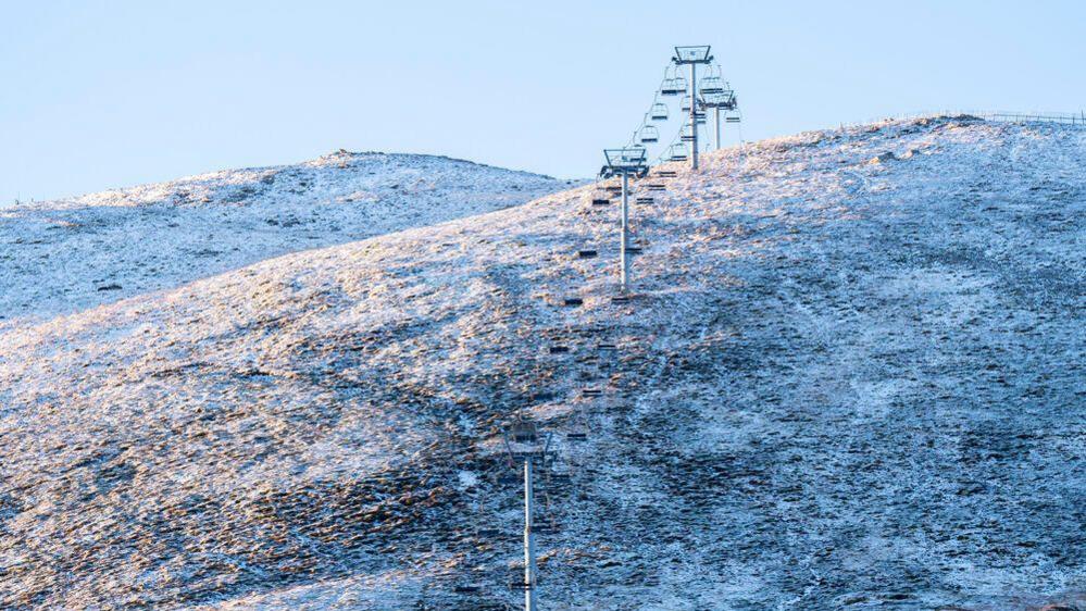 A hill covered in a light cover of snow with a cold white sky. A ski lift can be seen in the distance on the hill.