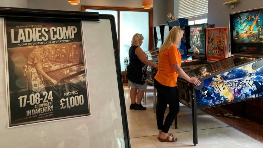 A woman in an orange top and another in a blue top play on Pinball machines - a notice in the foreground reads "Ladies Comp"