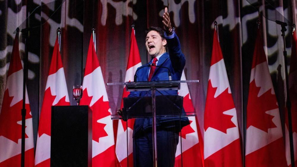 Canada’s Prime Minister Justin Trudeau speaks at a podium at the federal Liberal caucus holiday party, wearing a suit with a number of Canadian flags behind him