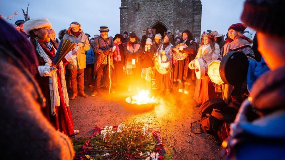 A crowd of people gathered around a fire pit, with a stone tower in the background. Some are carrying drums and there is a pile of flowers on the floor