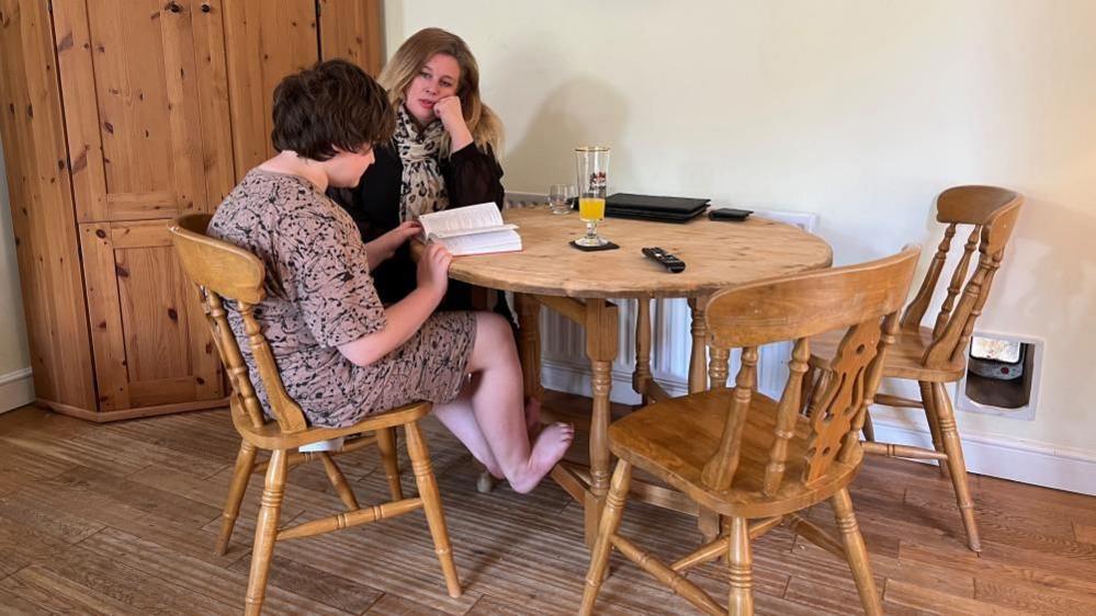 Sam and Suzi sitting at a table. Sam is reading a book.