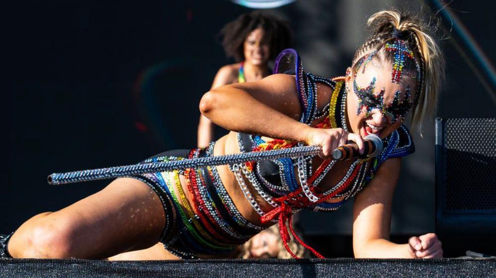 JoJo Siwa lying on a stage singing and performing at music festival Mighty Hoopla in London in 2024. She's wearing a black and brightly-coloured outfit covered in silver chains and holding a matching microphone. She has dark eye make-up which is surrounded by multi-coloured jewels.