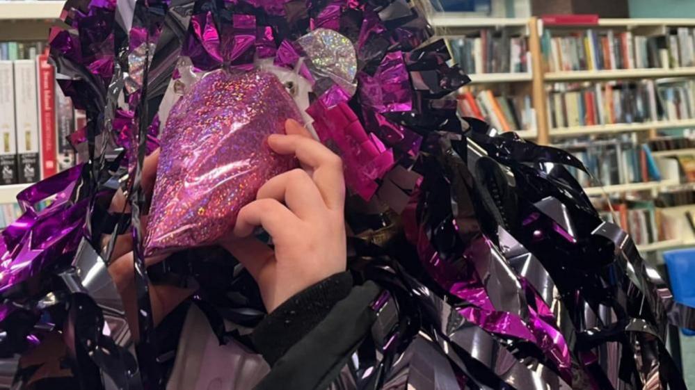 A crow costume made out of shiny black and purple material, standing in front of a row of books in a library