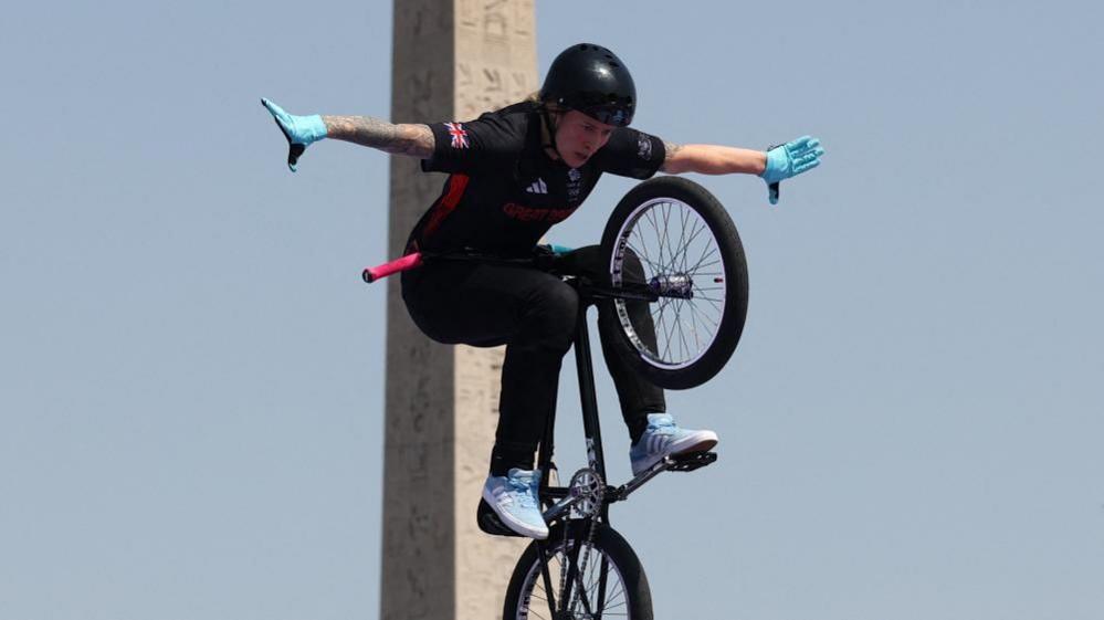 Charlotte Worthington with a black helmet and black shirt and trousers, with the Team GB logo on her sleeves. She is riding a black bike which is in the air and almost vertical alongside a stone pillar.