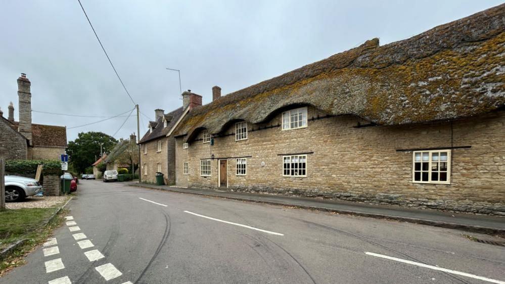 Village road junction showing large stone-built two-storey house with small windows. There are skid marks on the road.