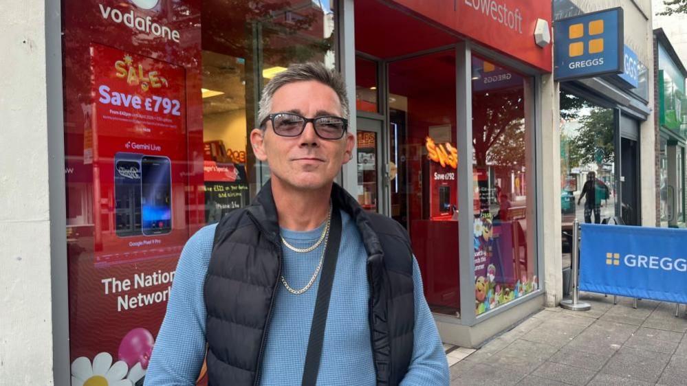 Tony McLeod is wearing a blue jersey, a black gilet and has a black bag strap over his shoulder. He also has two chains around his neck and is wearing tinted glasses. He is clean-shaven and has medium-length brown hair, greying at the sides. He is standing outside the Vodafone shop in Lowestoft.