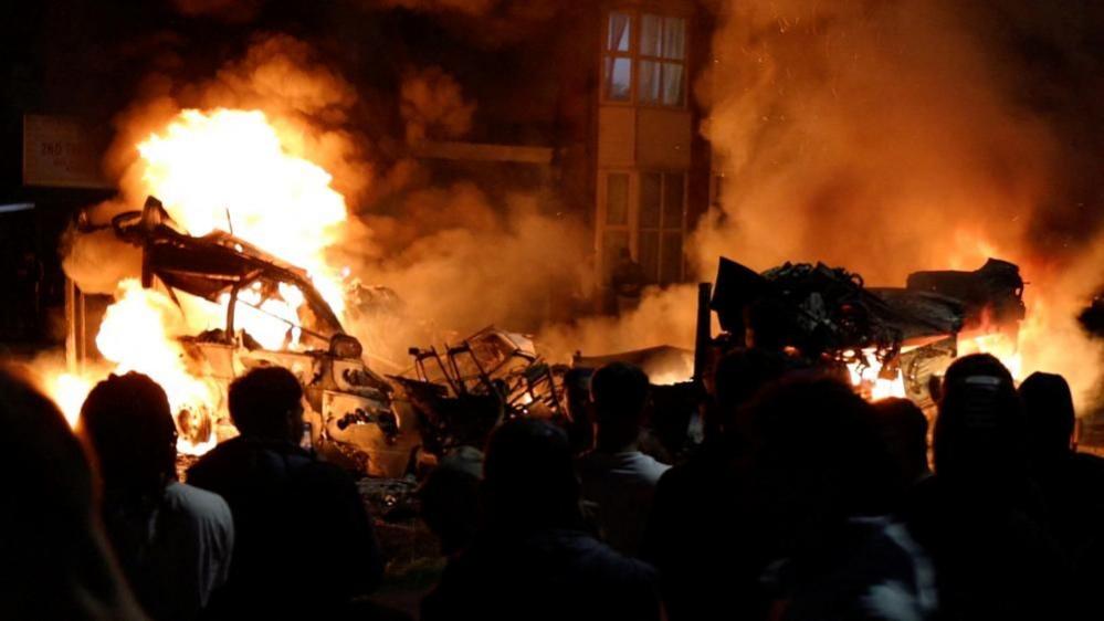 Crowds stand in front of a burning double decker bus with flames leaping into the night sky