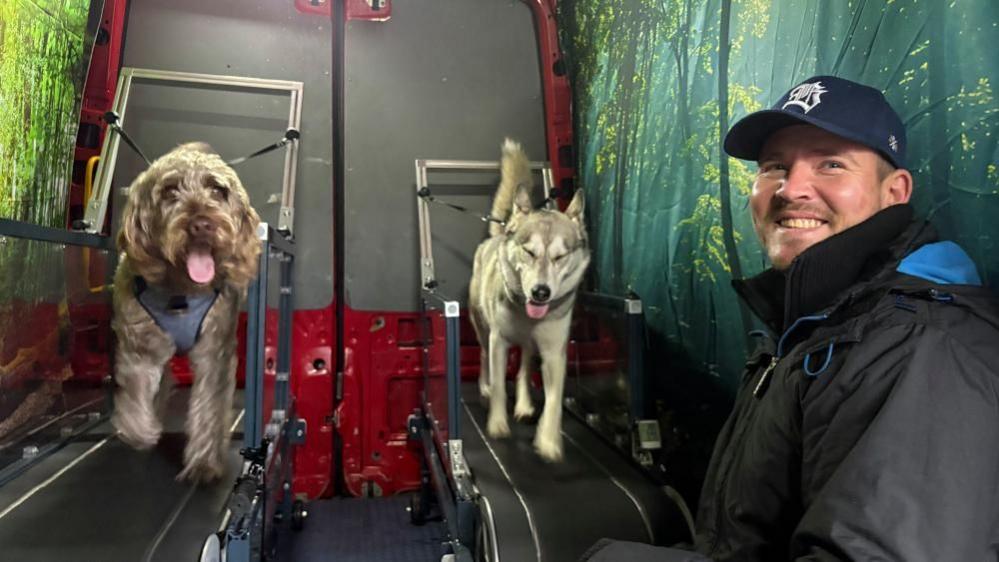 A man wearing a black rain coat and a baseball cap smiles at the camera. Behind him are two dogs running on two treadmills. The dog on the right is a grey husky type and the one on the left is a caramel coloured Poodle cross Labrador. They are all inside a red transit van, with large forest images on the inside walls.