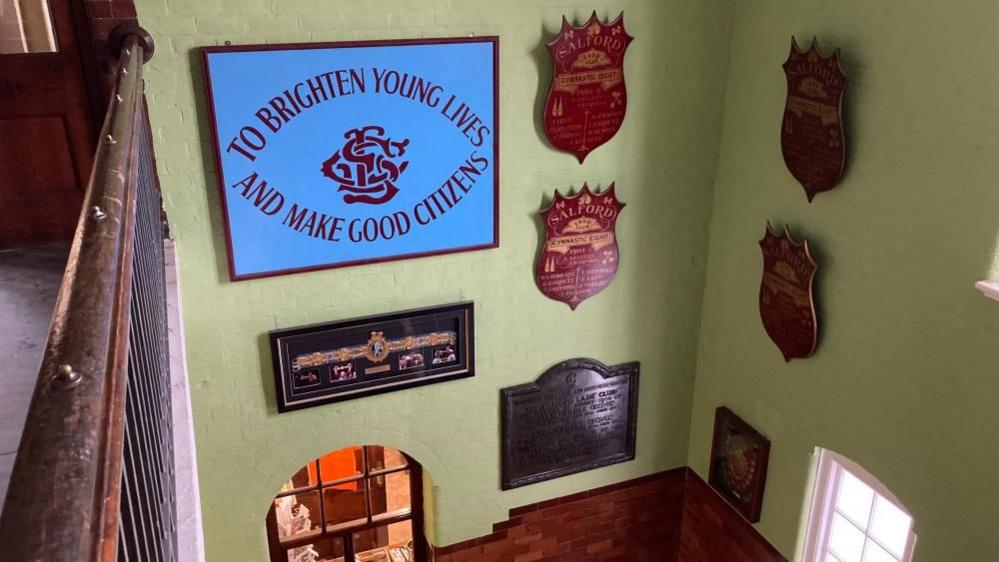 A number of crests and a sign that reads 'To brighten young lives and make good citizens' adorn the tiled walls of the Salford Lads Club. 