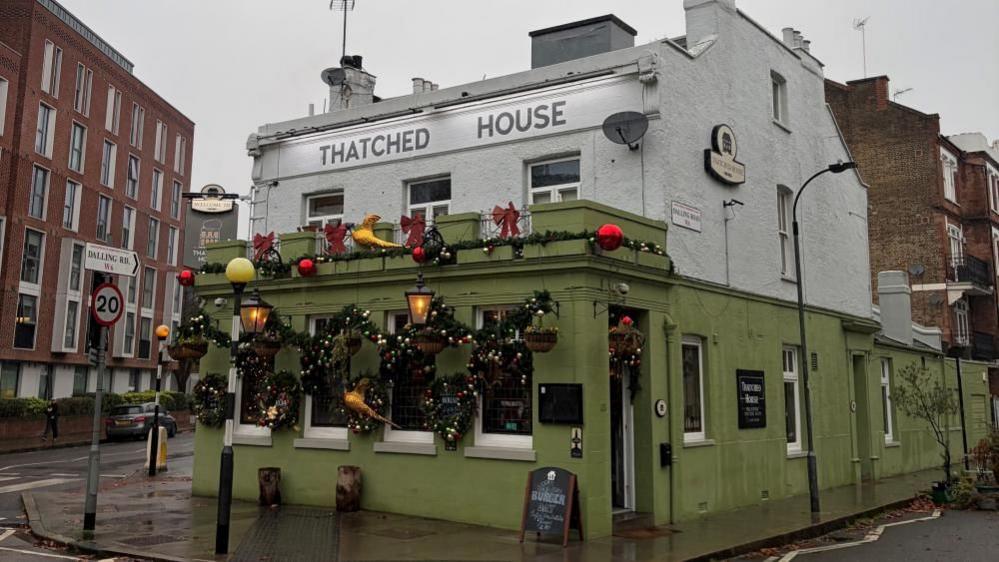 A pub with a green brick first floor and white top floor. Signage reads "Thatched House and the windows are decorated with festive garlands and baubles.