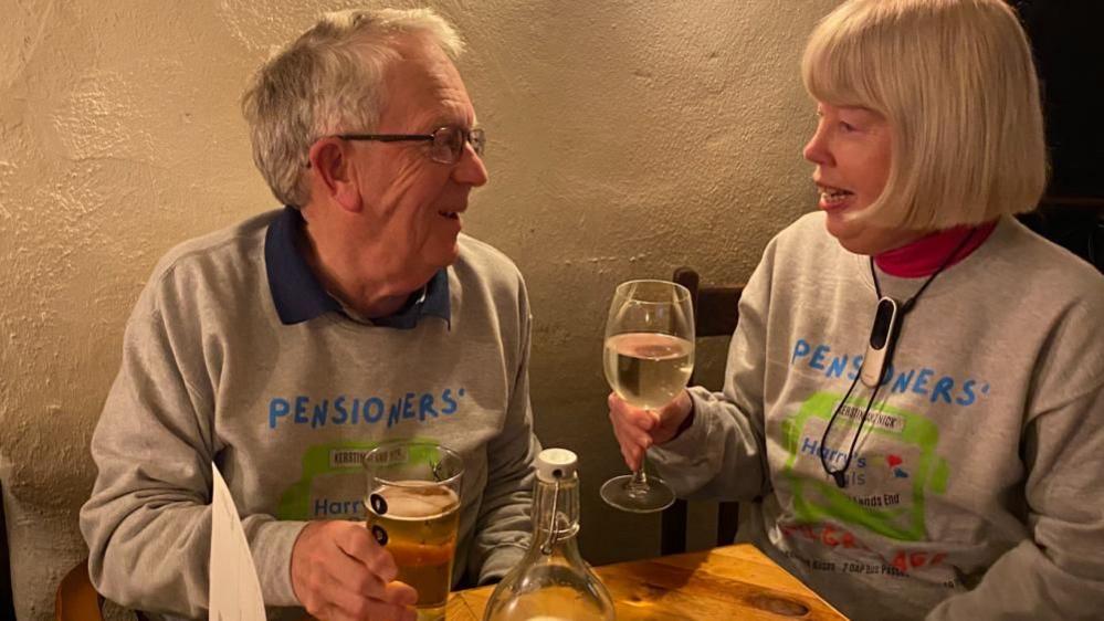 Mr and Mrs Banham in their grey sweatshirts sitting at a table in a pub. Mrs Banham has a glass of wine in her hand and Mr Banham is holding a pint of beer.