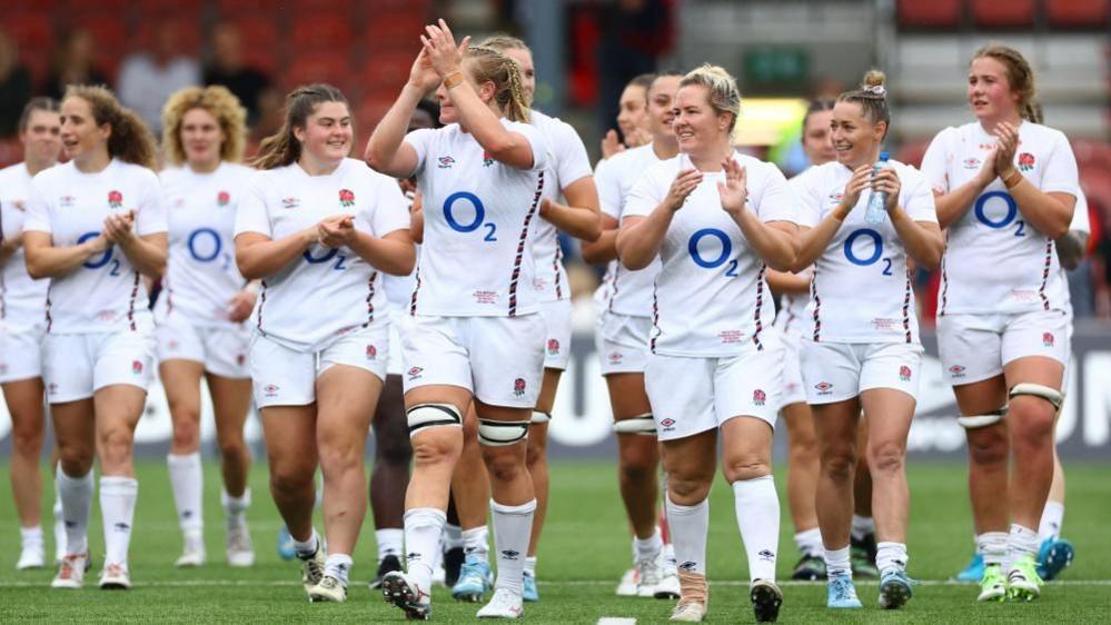 The England Women's team line up on the grass at a rugby ground.  They are all wearing white shirts and shorts with England and O2 logos. Some are clapping. Advertising hoardings and some spectators are visible behind them.