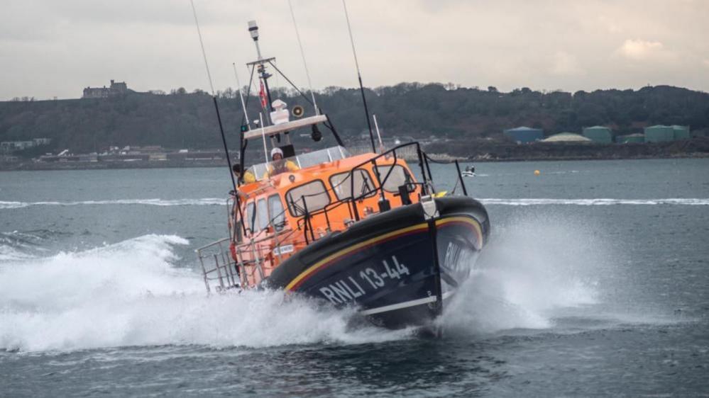 The RNLI Lifeboat fleet numbered 13-44, the George and Frances Phelon, under power with white sea spray either side of the hull. Two crew members can be seen in yellow waterproof clothing on the fly bridge of the vessel. In the backdrop is a series of silos and buildings, with cliffs and trees visible.