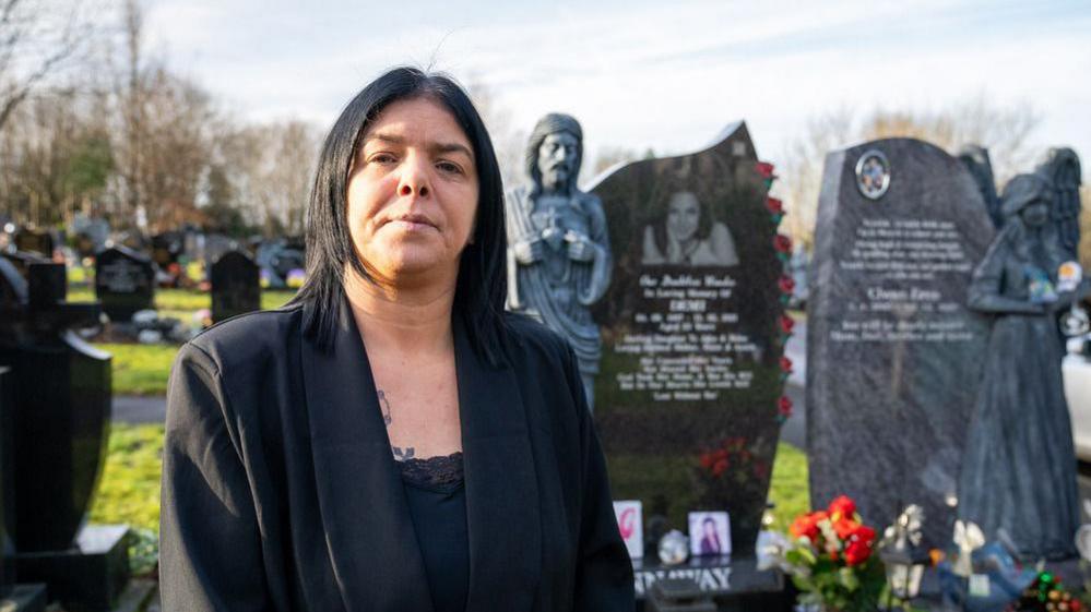 Helen stands in front of her daughter's grave. She is dressed in black and looks at the camera. 