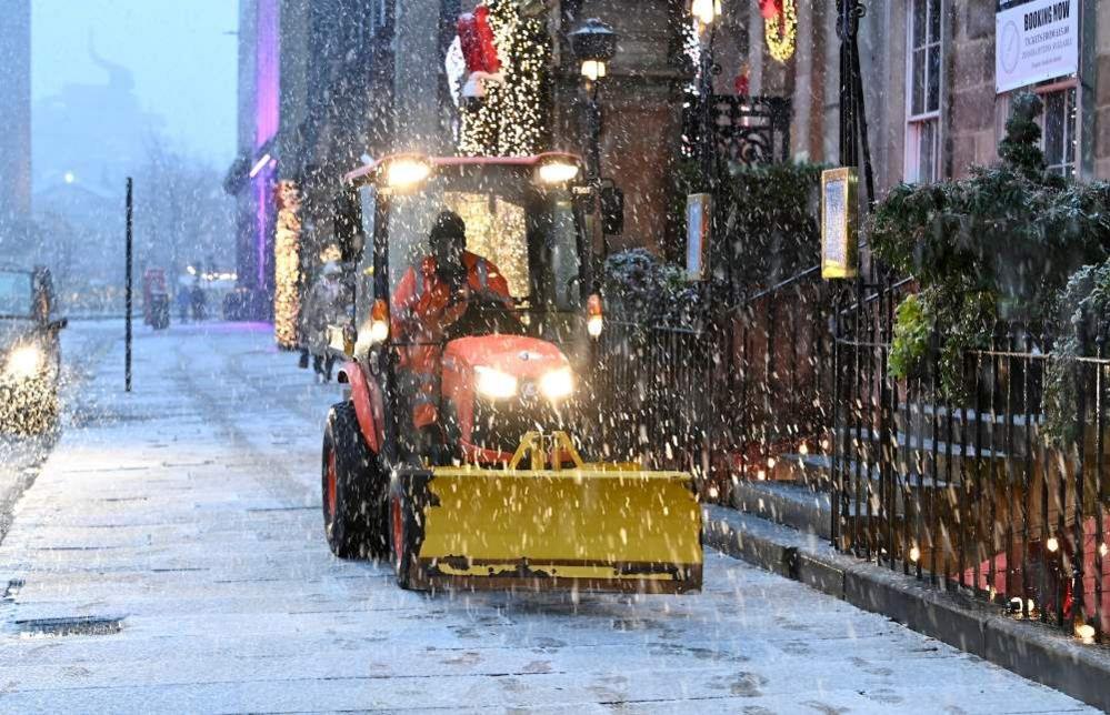 A man wearing a hi-vis taking driving orange and yellow snow plow with lights on along a snow covered pavement.