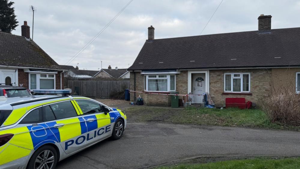 A police vehicle outside a bungalow on Grounds Avenue, March. The house has two windows, a door and tape across the front of the house. Another bungalow is to the right, with one window and a door in the shot. There is a drive that goes in front of the house. 