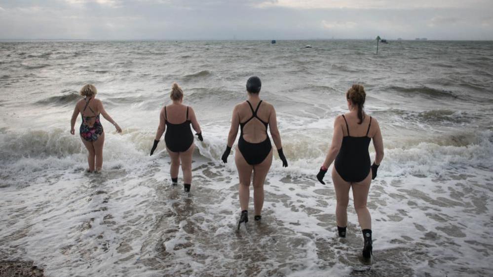 Four swimmers in black wetsuits and gloves walk into the sea as the waves crash in. The sky is grey.