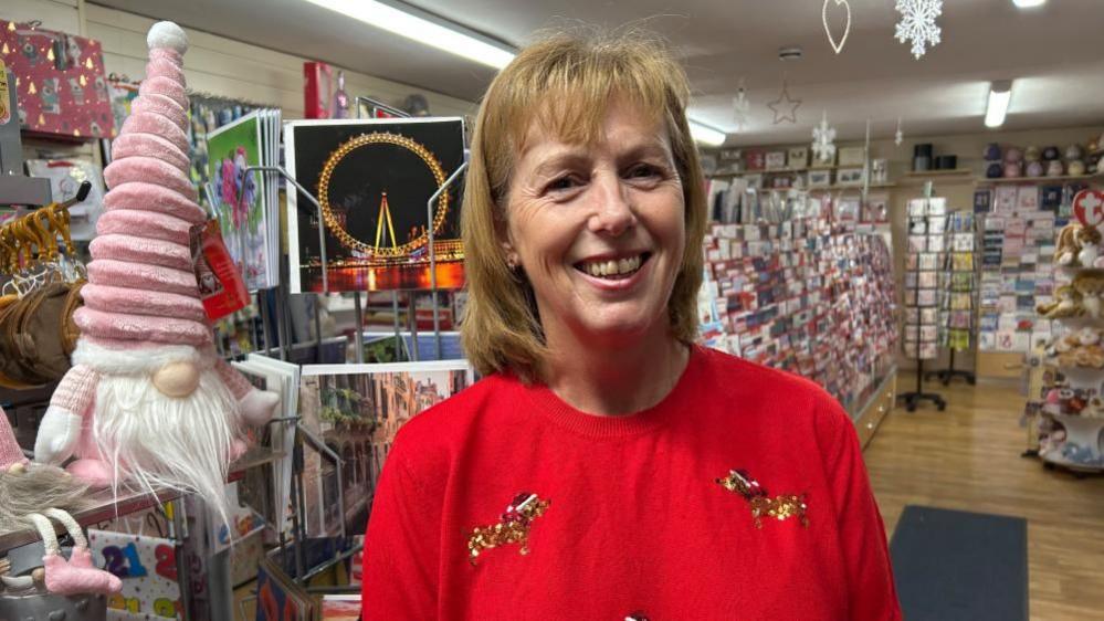 Anita Andrews in a red jumper with shoulder-length auburn hair. She is stood in her card shop with hundreds of cards behind her. To her left is a pink soft Father Christmas toy.