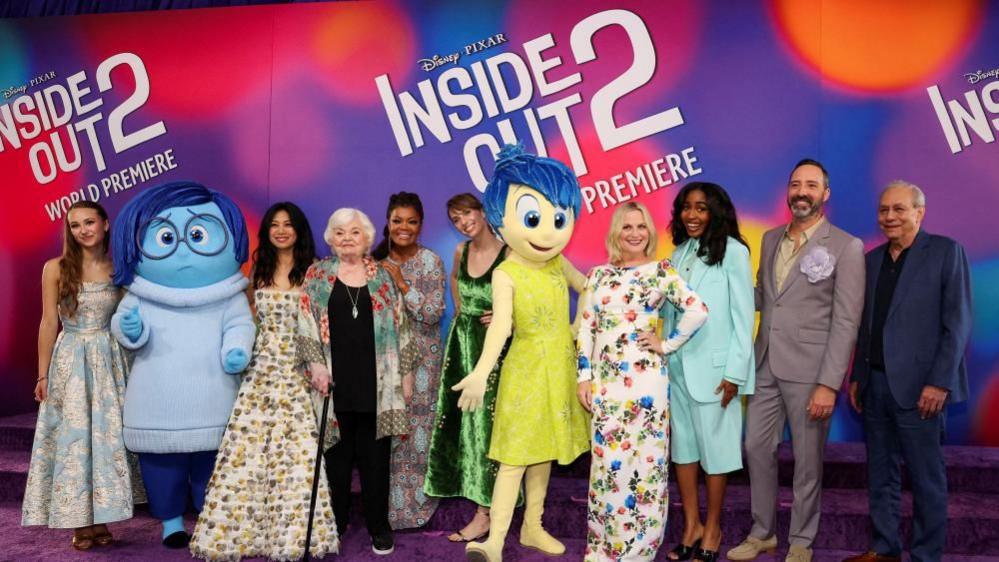 The cast of Inside Out 2 with two people dressed as two of the characters in front of a sign which says "Inside Out 2"