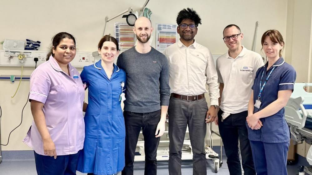 Three women and three men stand smiling in a hospital ward. The women are wearing nurse uniforms.