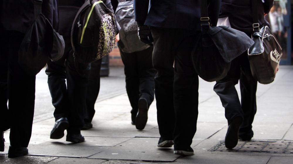 A group of schoolchildren walking along the pavement with their school bags over their shoulders. 