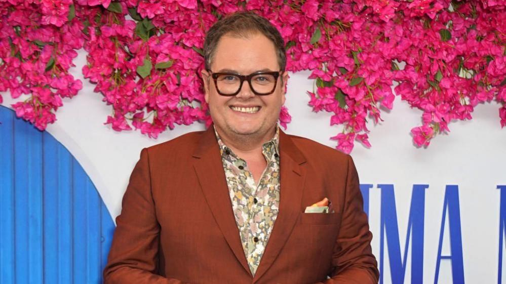 Alan Carr wearing a brown suit and floral shirt