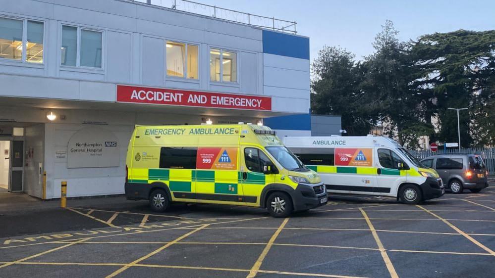 The entrance to Northampton General A&E department, showing red "accident and emergency" sign above the white entrance. There is a yellow ambulance and a white ambulance parked by the entrance.