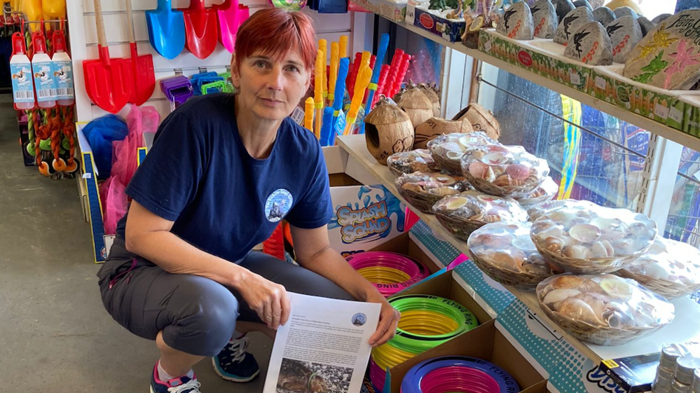  Clair Murgatroyd in a seaside shop crouching next to a display of plastic toys, she holds a printed newsletter with a photo of a dead seal. 