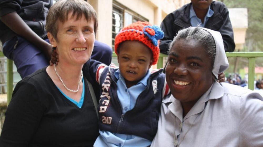 Gena, left, is smiling at the camera. She has short brown hair and is wearing a black long sleeve top. A child is in the middle, wearing a bright red hat and a navy zip up jacket. Another woman, right, is smiling at the camera. She is wearing a light grey dress and has a matching hair scarf on.