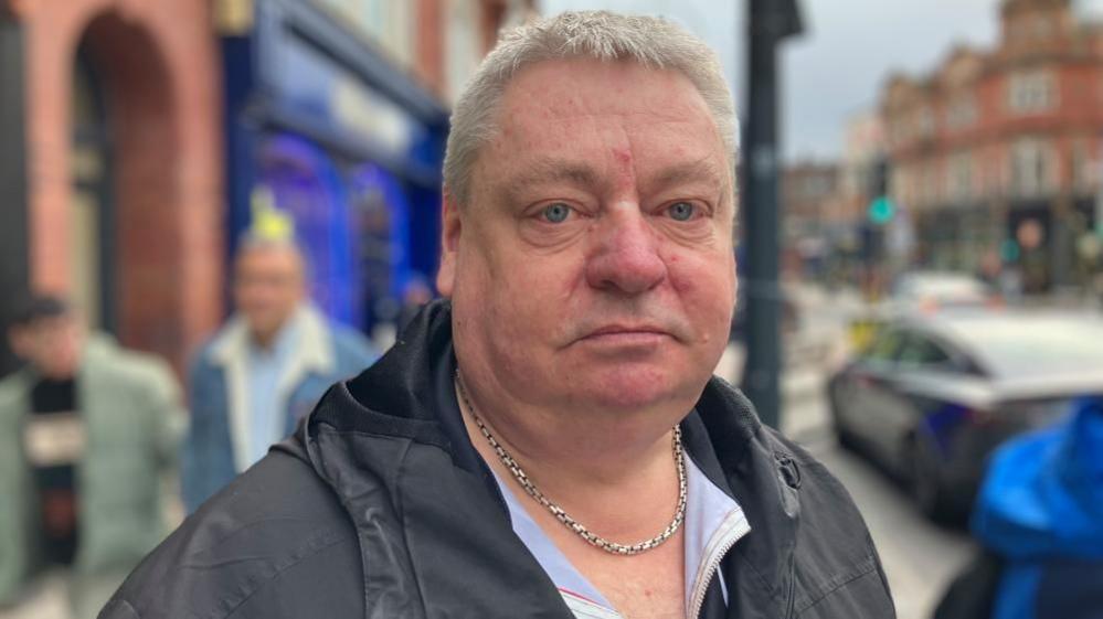 A middle-aged man stands in a busy street with a stern expression. He has blue eyes and greying hair and wears a silver chain under a shirt and black coat.