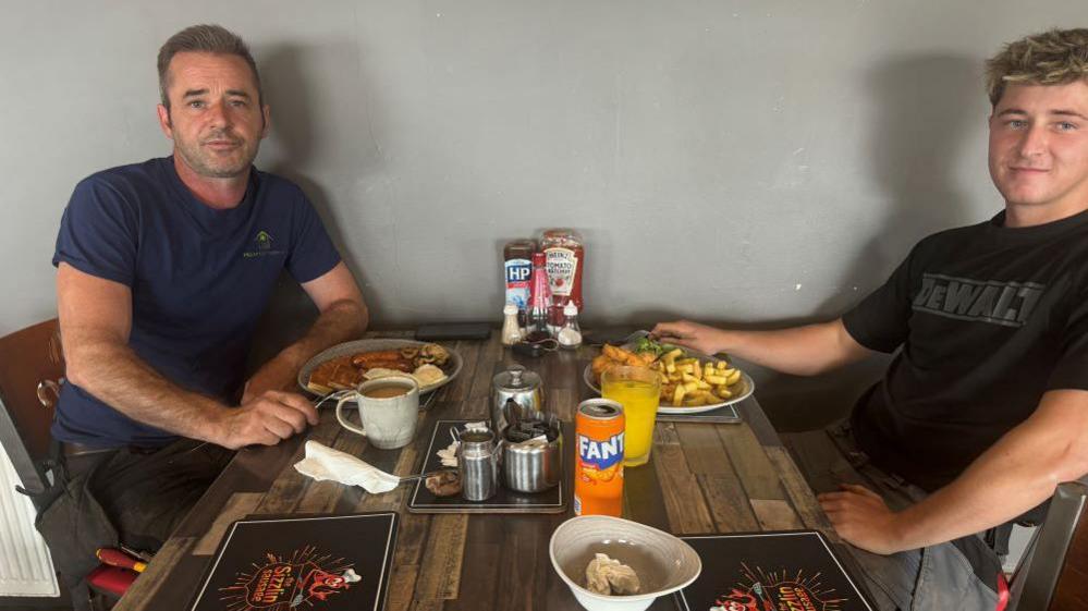 Paul wearing a navy t-shirt and black work trousers and tool belt sat at table with colleague also in workwear. They are sat a table with a fry up in front of Paul and chips in front of his colleague.  