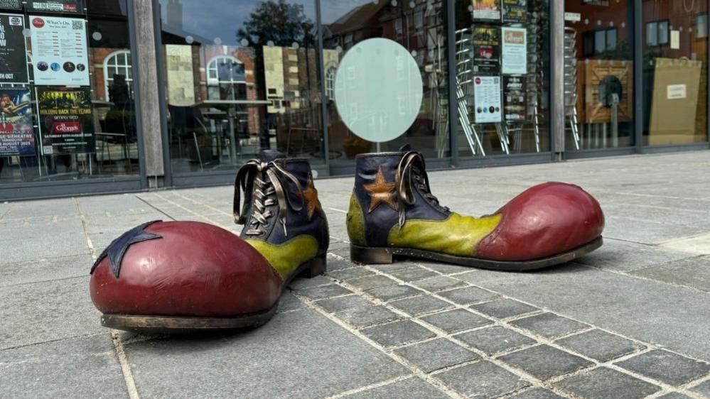Two giant clown shoes, painted in black uppers, with yellow mid banding, and red enlarged toecaps, stands on a stainless steel frame that will be buried underground. In the backdrop is St George's Theatre Pavilion.
