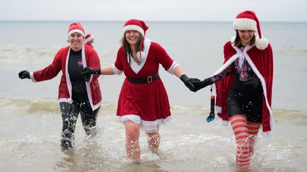 Three women dressed as santa claus holding hands step out of the sea