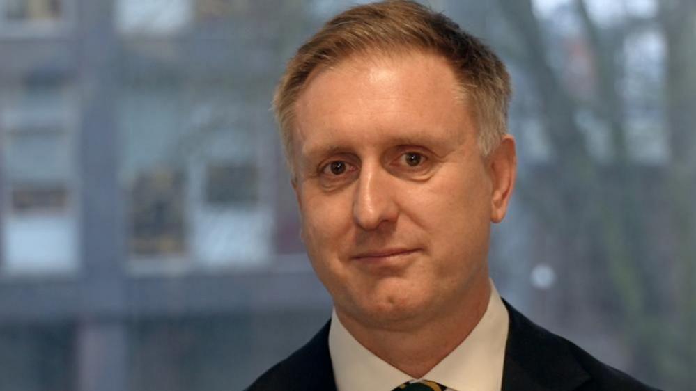 Dr Ed Garratt is smiling at the camera and is wearing a suit. He has short brown hair, going grey at the sides. The background is blurred it but looks like he is standing in front of a window.