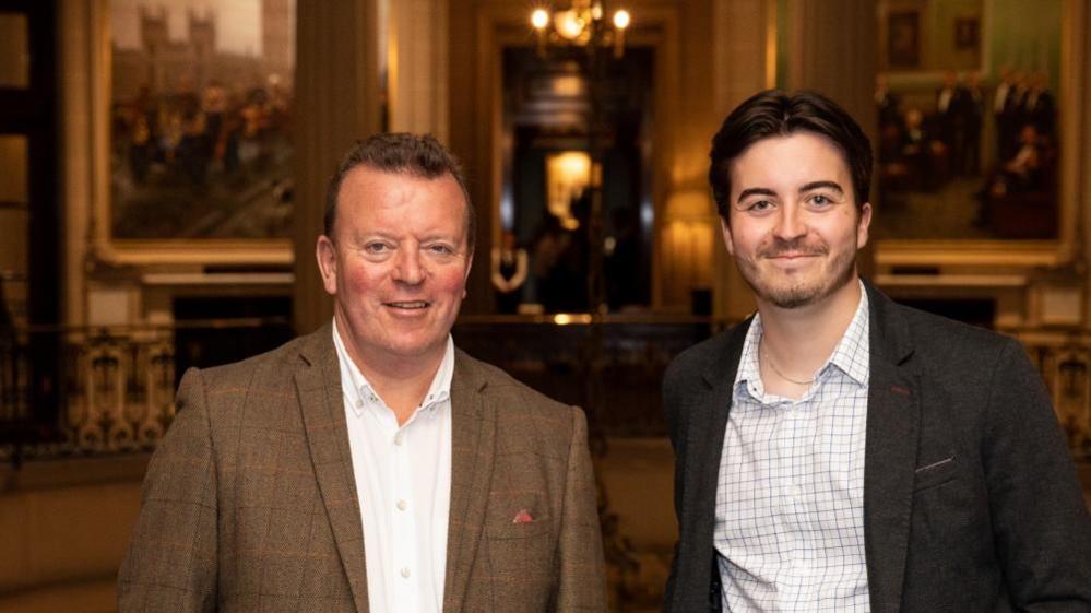 Nigel McNally with short brown hair wearing a brown jacket and Joel with dark hair and dark brown jacket